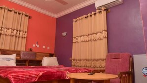 A room with two beds, a round table, a chair, an air conditioning unit, and a wall-mounted light fixture.