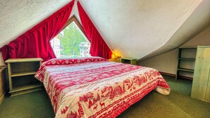 A cozy attic bedroom with a sloped ceiling, a large bed with a red and white patterned bedspread, a nightstand, and a window with red curtains.