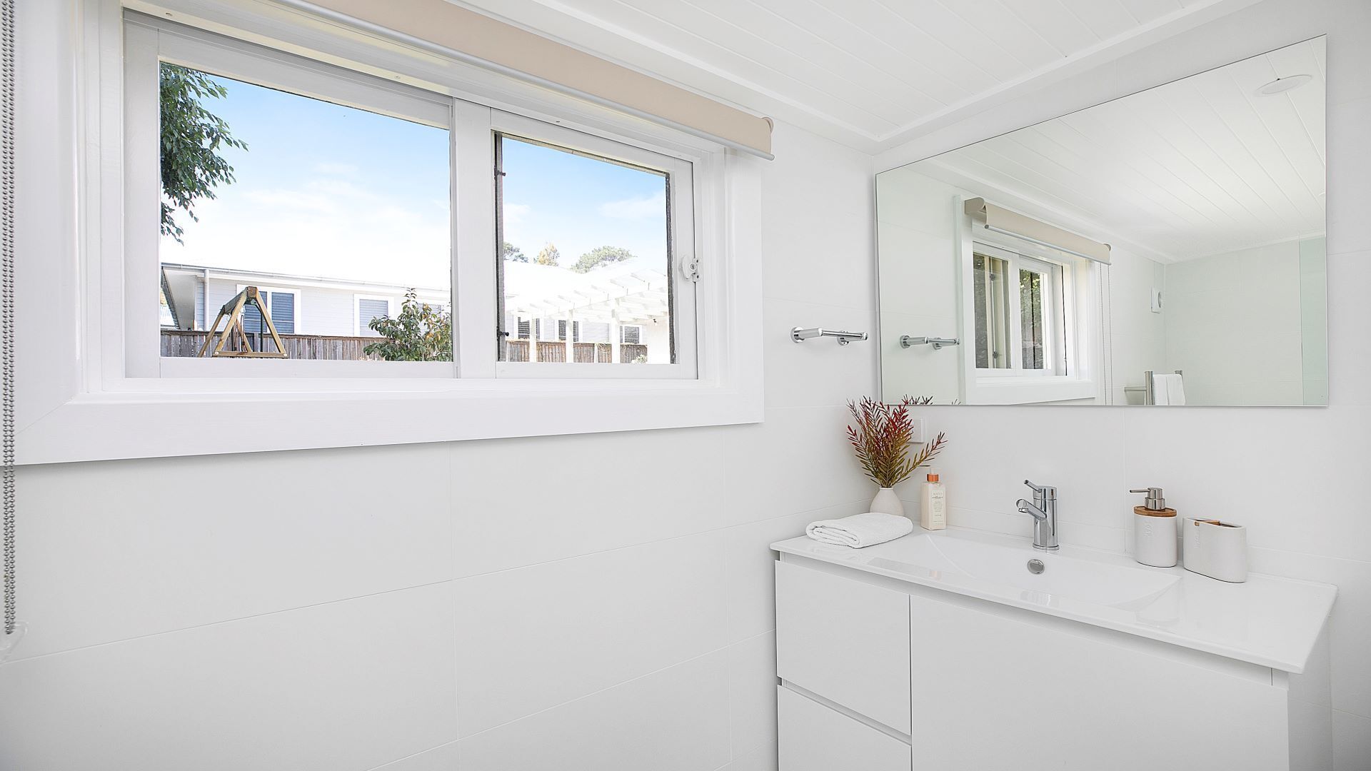 A modern bathroom with a large window, a white vanity, a mirror, and a view of a residential area.