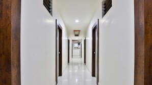 A long, narrow hallway with white walls and wooden doors, leading to a room with a ceiling light. - Hotel Bansidhar (Veraval)