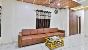 A living room with a brown leather sofa, a wooden coffee table, a ceiling fan, and a patterned curtain. - Hotel Bansidhar (Veraval)
