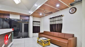 A modern living room with a brown leather sofa, a wooden coffee table, and a reception desk with a glass partition. - Hotel Bansidhar (Veraval)