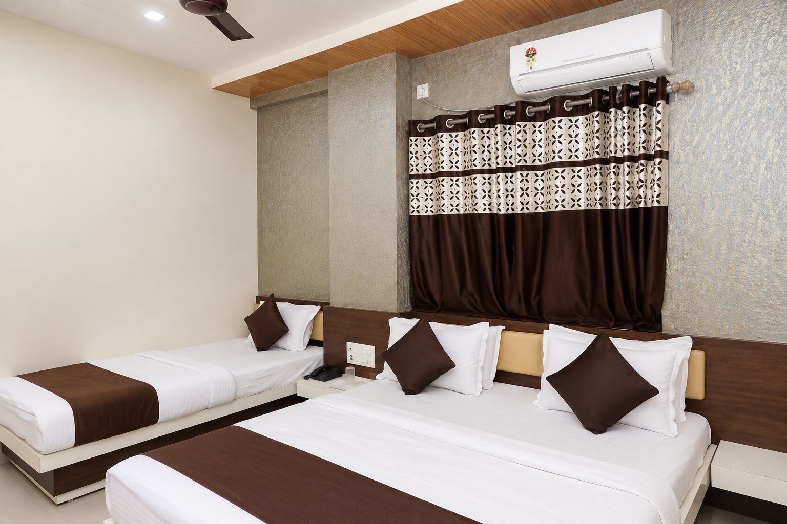 A hotel room with two beds, a ceiling fan, an air conditioning unit, and a patterned curtain.