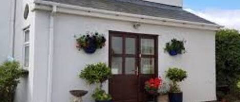 Un edificio blanco con puerta marrón, ventanas y un pequeño porche con plantas en macetas.