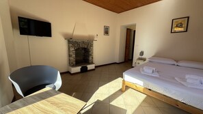 A bedroom with a stone fireplace, a bed with white linens, a desk with a chair, and a television on the wall.