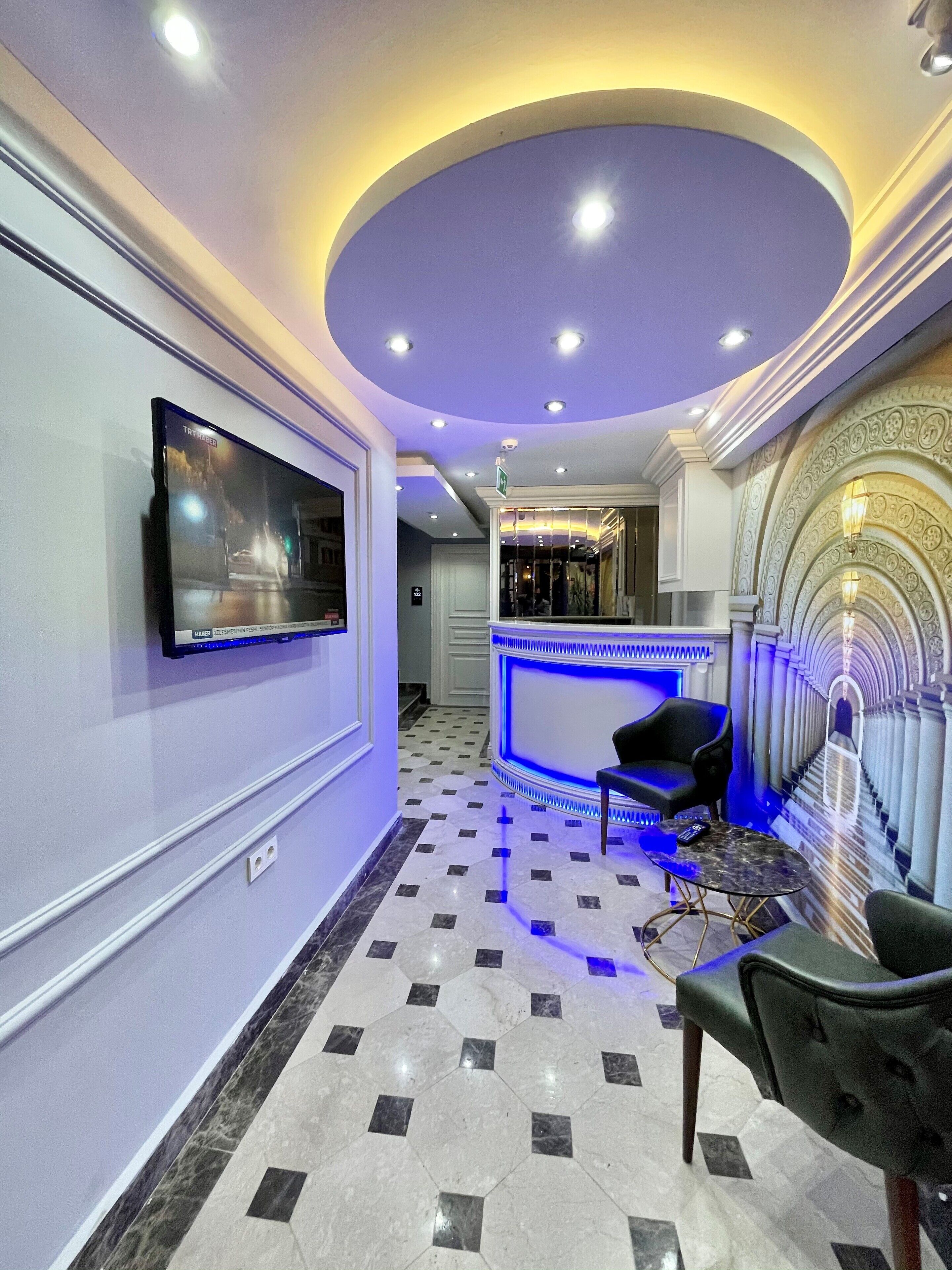Vestíbulo de hotel con mostrador de recepción, un televisor de pantalla plana en la pared, una silla de cuero negro y un suelo con diseño de mármol.