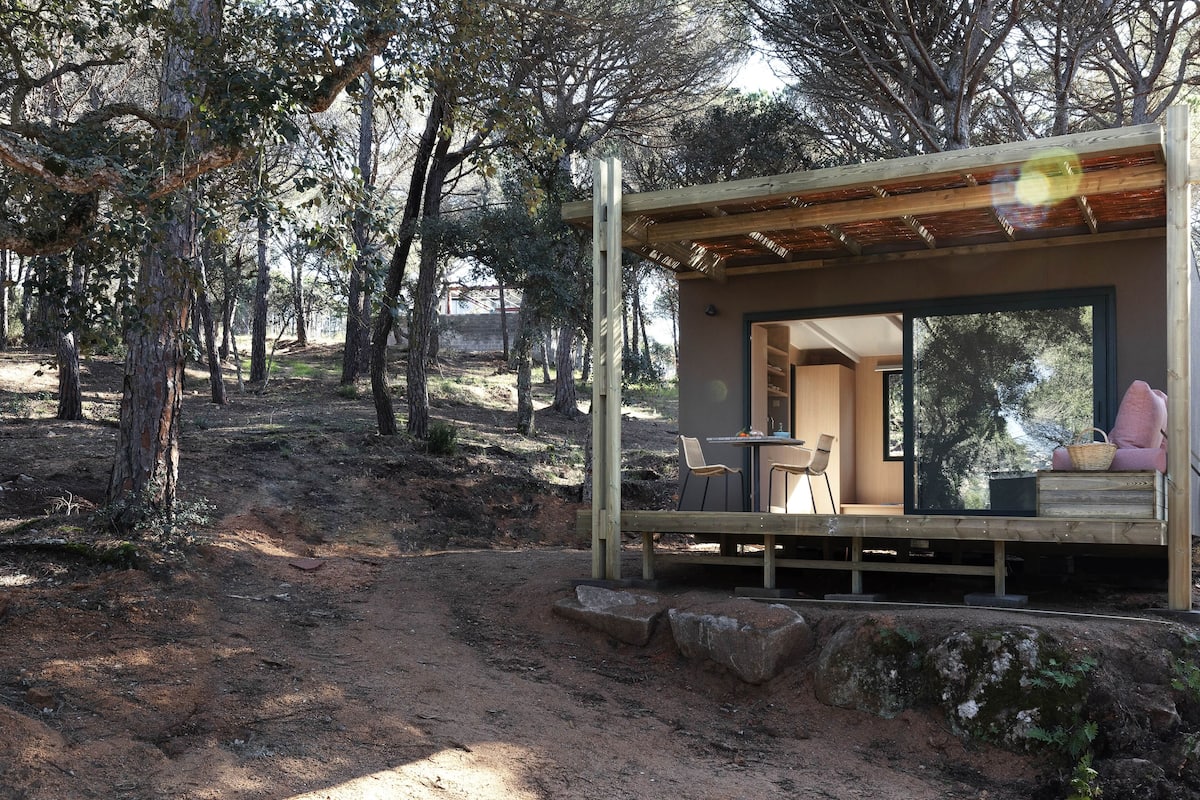 Una cabaña rústica con terraza de madera, un comedor con sillas y un amplio ventanal con vistas al bosque circundante.