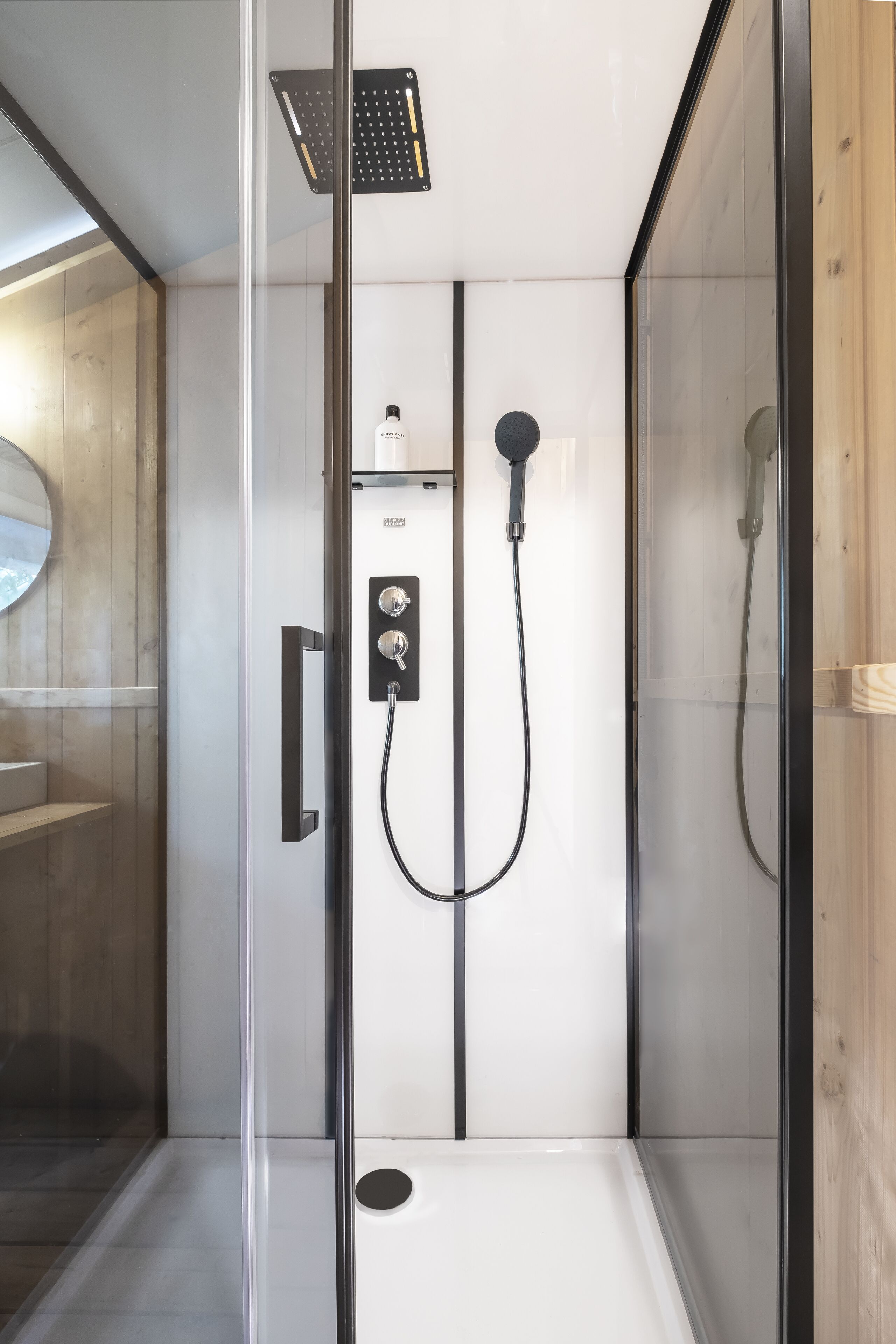 A modern shower enclosure with a glass door, a handheld showerhead, a wall-mounted control panel, and a small shelf with toiletries.