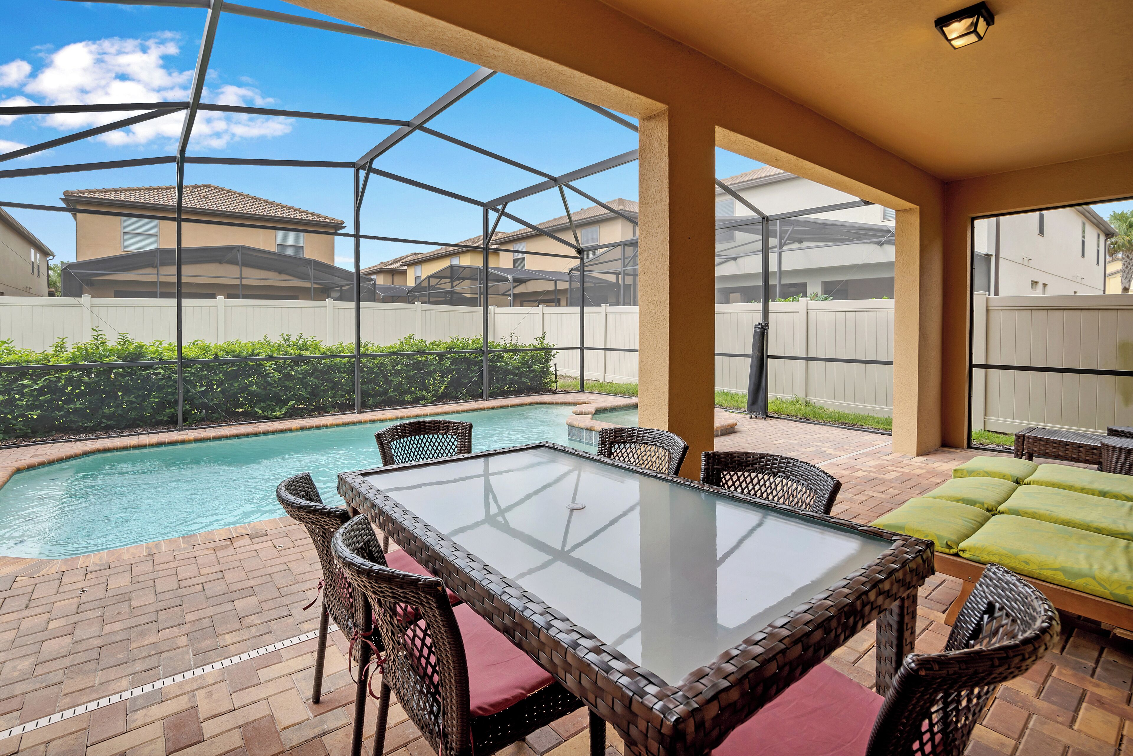 A covered outdoor area with a glass-top table and wicker chairs, a pool, and a view of neighboring houses.