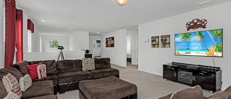 A living room with a sectional sofa, a flat-screen TV mounted on the wall, and a kitchenette area.