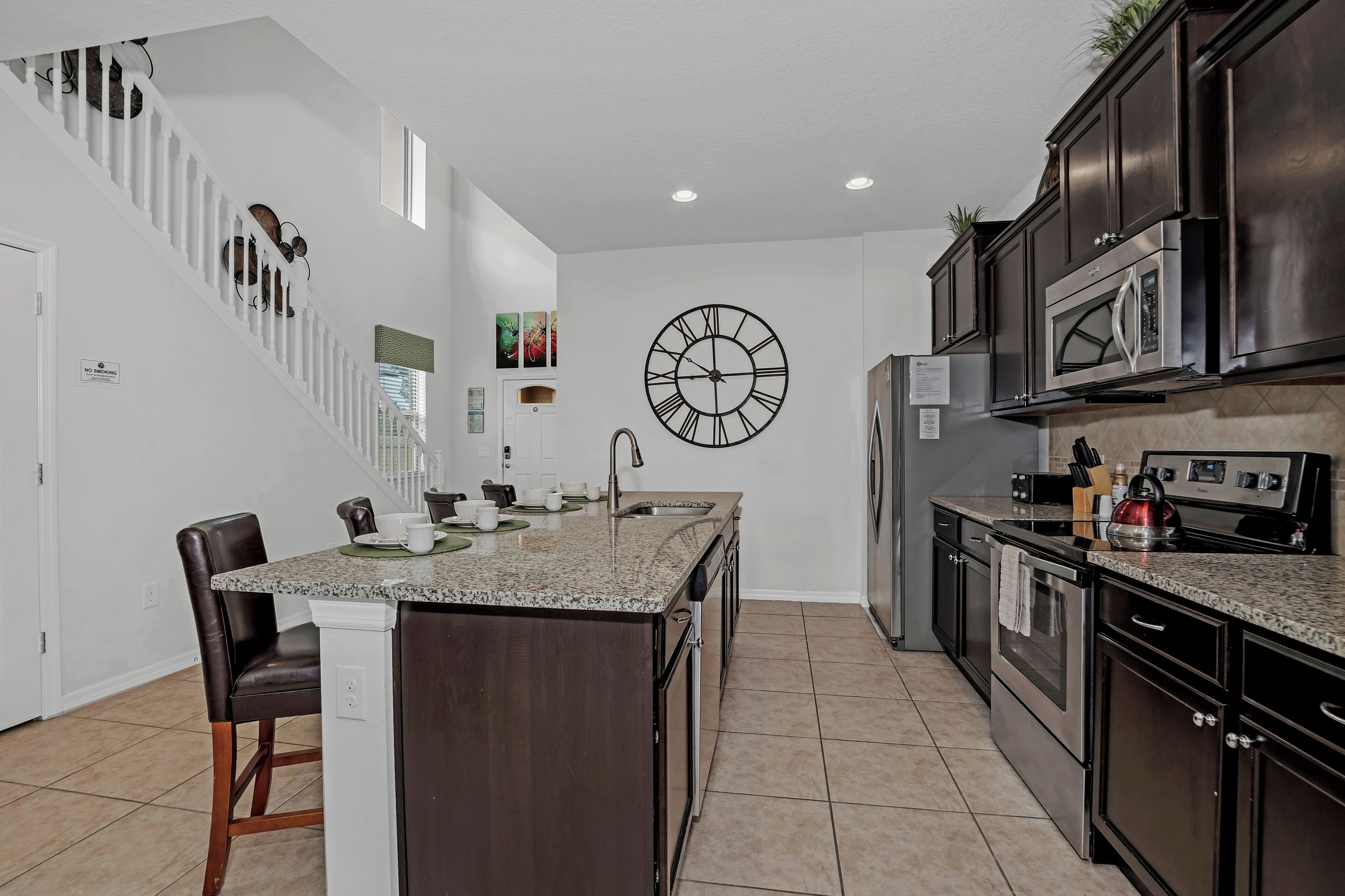 A modern kitchen with a central island, dark cabinetry, stainless steel appliances, and a large wall clock.