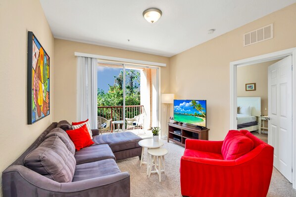 A living room with a grey sofa, red armchair, and a TV. There's a balcony with a view of trees and a white door.