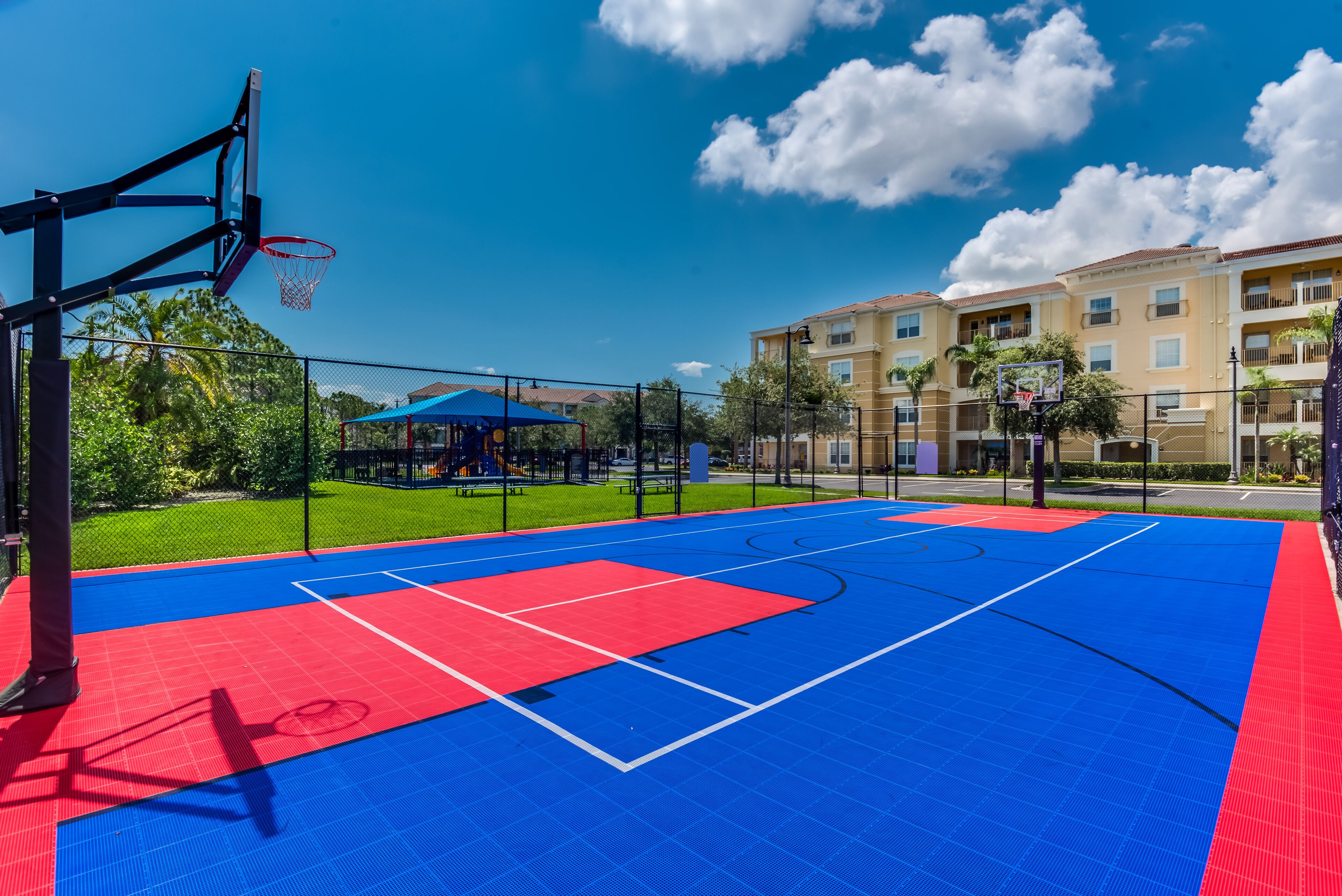 Een basketbalveld buiten met een blauwe en rode speeloppervlakte, omheind door een hek en met een gebouw op de achtergrond.