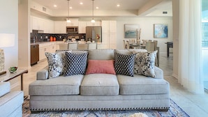 A modern living room with a grey sofa, patterned cushions, a dining area, and a kitchen in the background. - Modern Condo Near Disney Foosball Water Park! (Kissimmee)