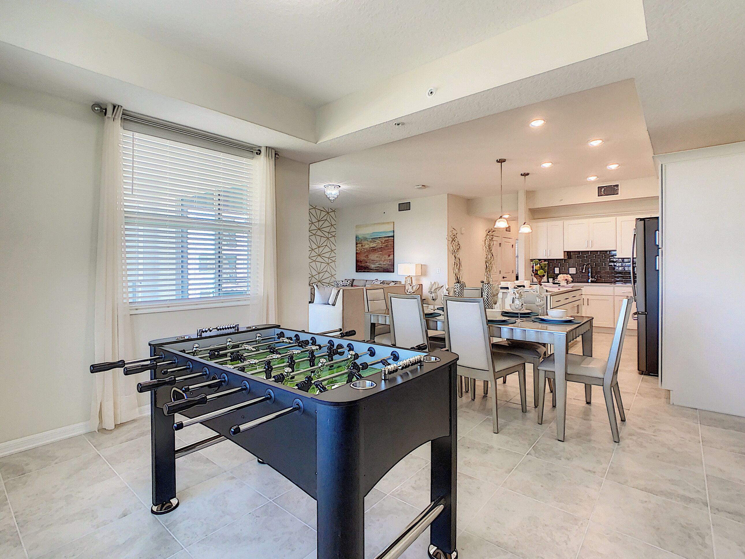 A modern living room with a foosball table, a dining area with a chandelier, and a kitchen with dark countertops.