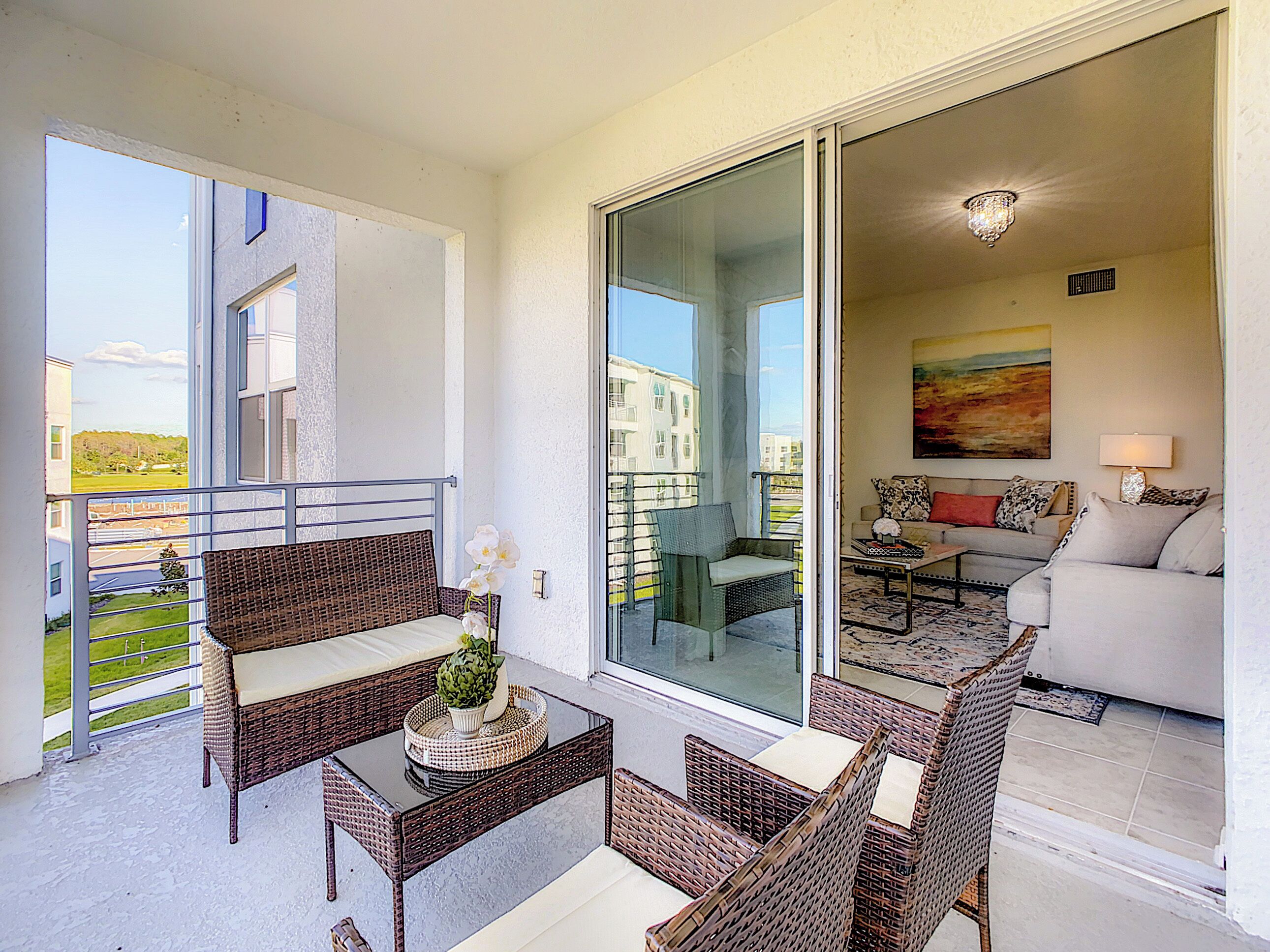 Een balkon met rieten meubilair, een glazen deur naar een woonkamer met een bank en een schilderij.