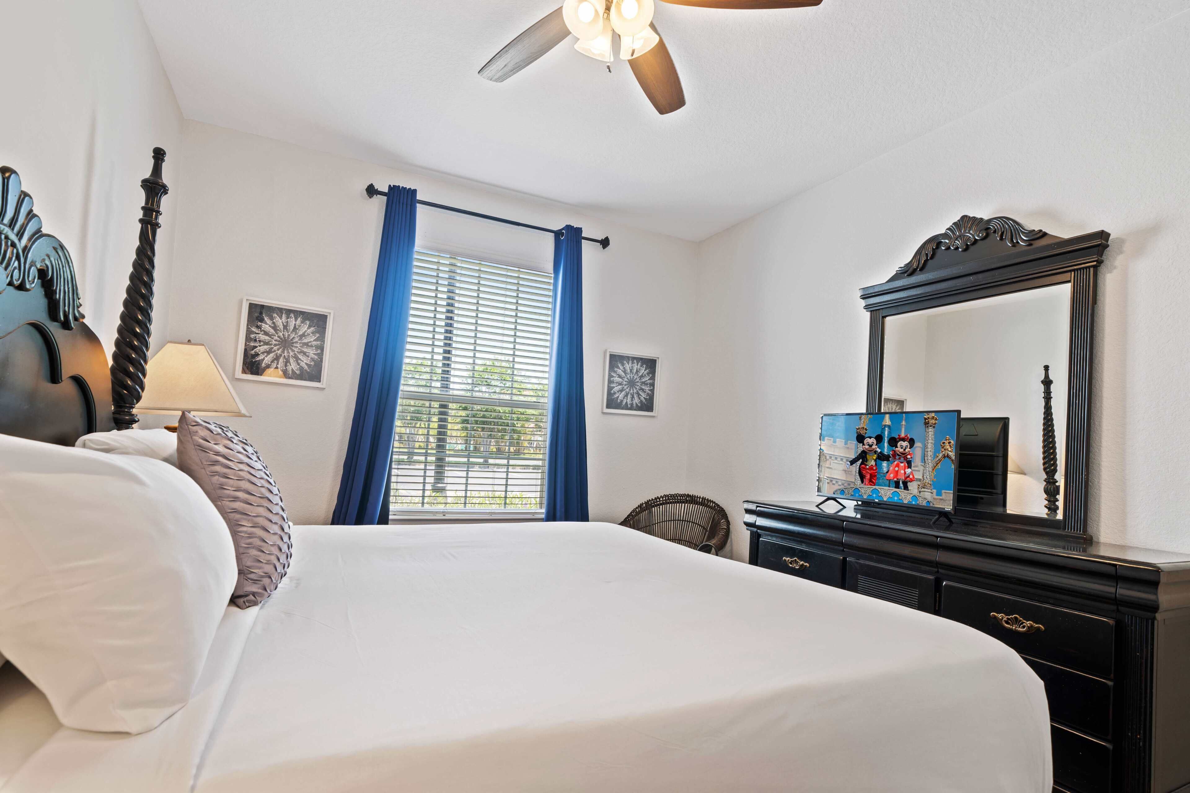 A hotel room with a bed, a ceiling fan, a window with blue curtains, a dresser with a mirror, and a lamp.
