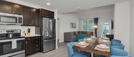 A modern kitchen with dark cabinets, stainless steel appliances, and a dining area with blue chairs and a wooden table.