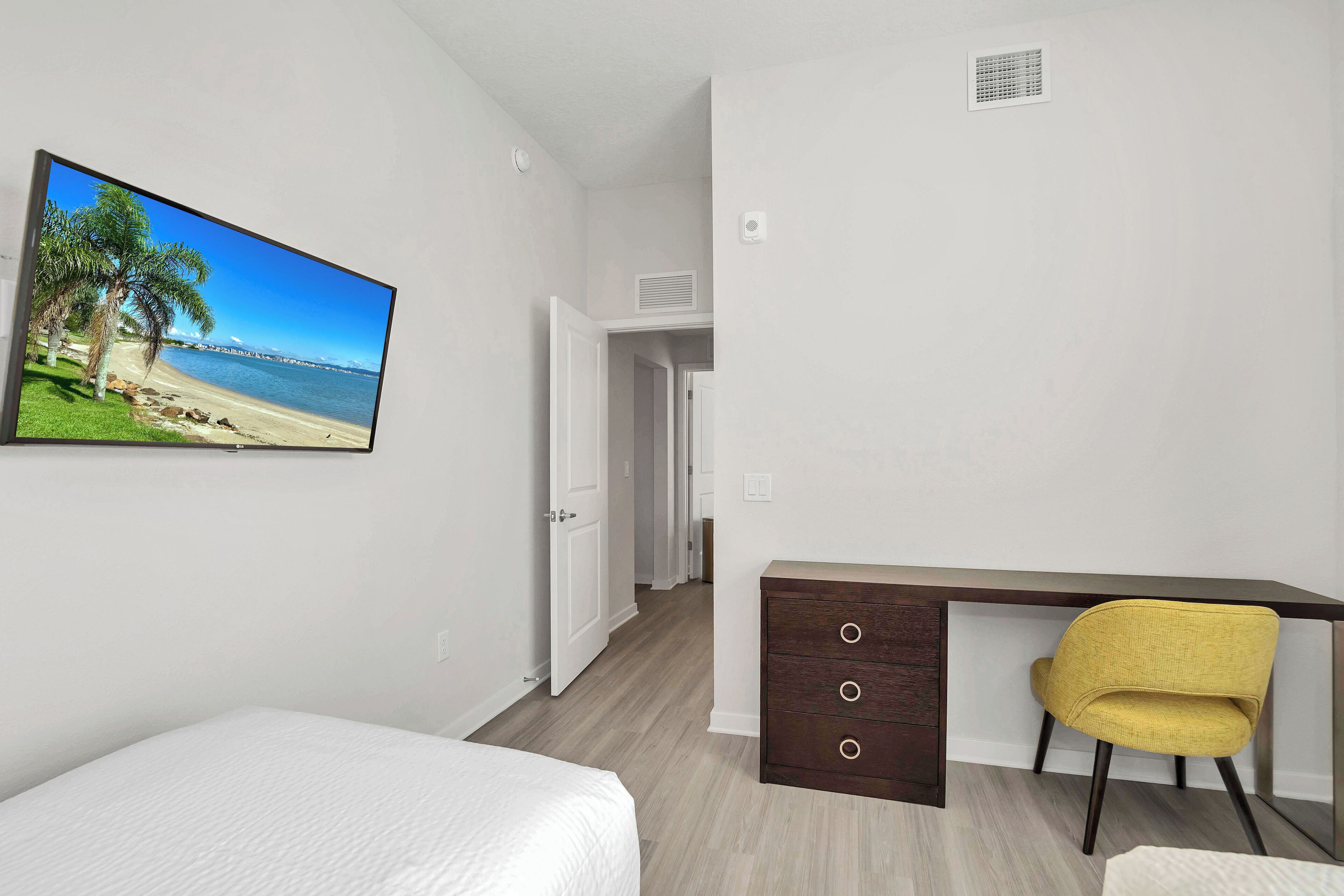 Une chambre à coucher avec un lit, un bureau et une chaise, un téléviseur diffusant une scène de plage, et une porte donnant sur une autre pièce.