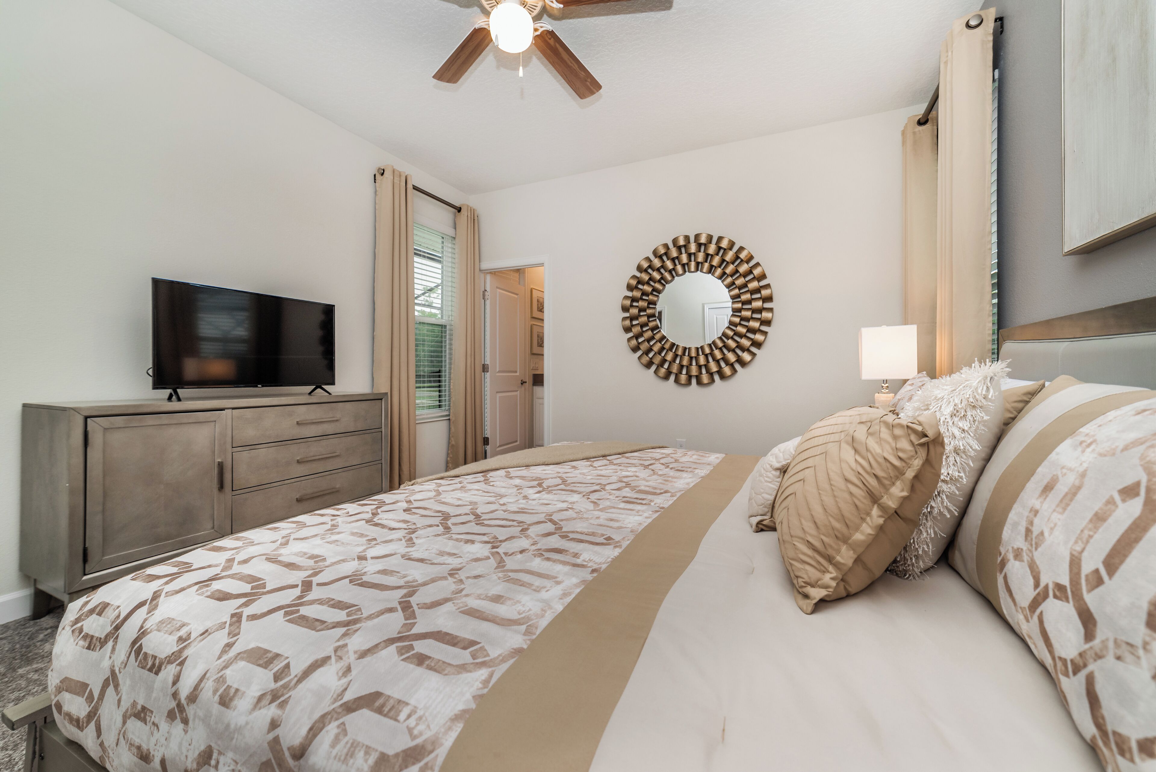 Un dormitorio con cama, televisión, ventilador de techo, espejo y ventana con cortinas.