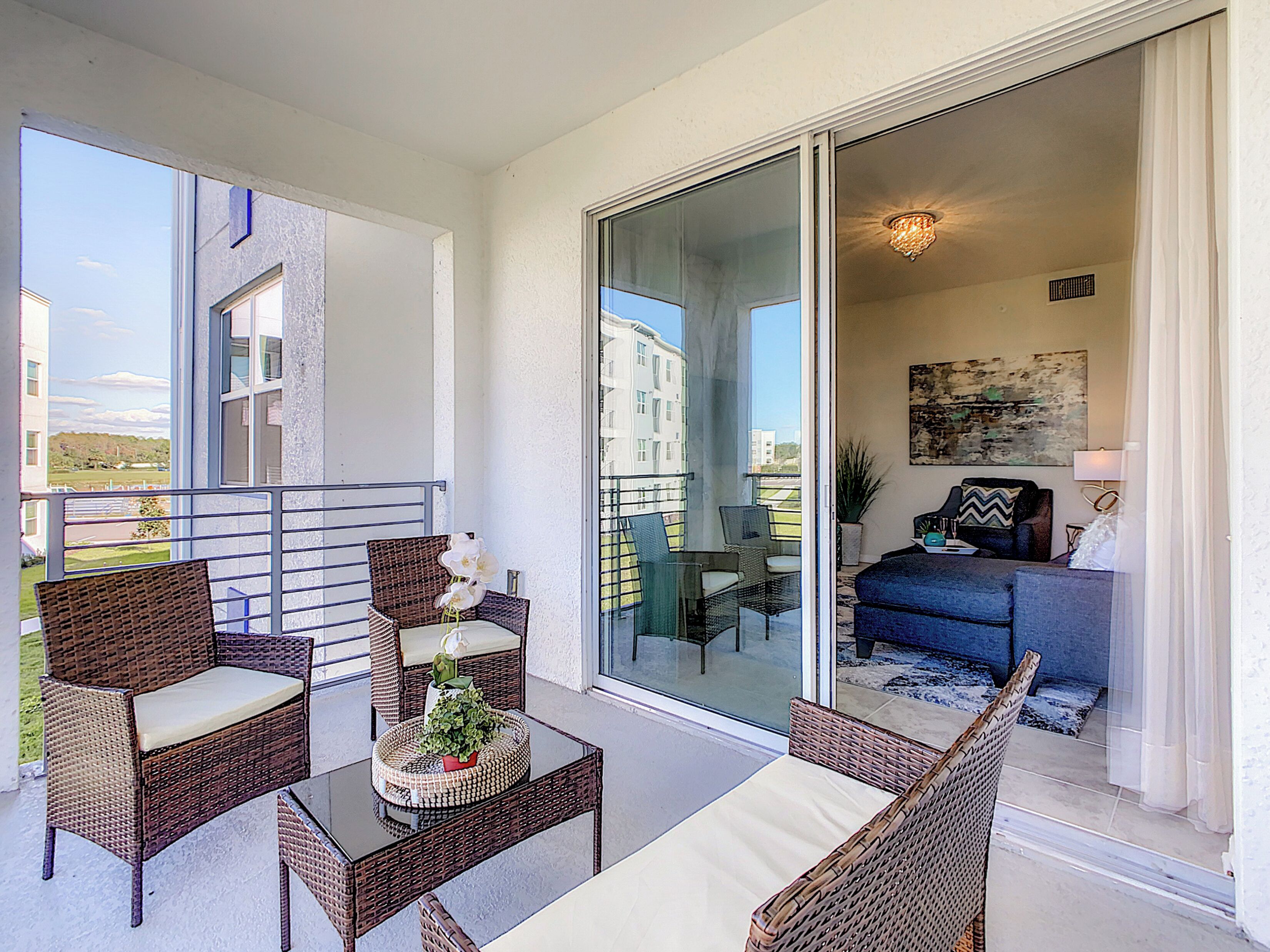 A balcony with wicker furniture, a glass door leading to a living room, and a view of buildings in the background.