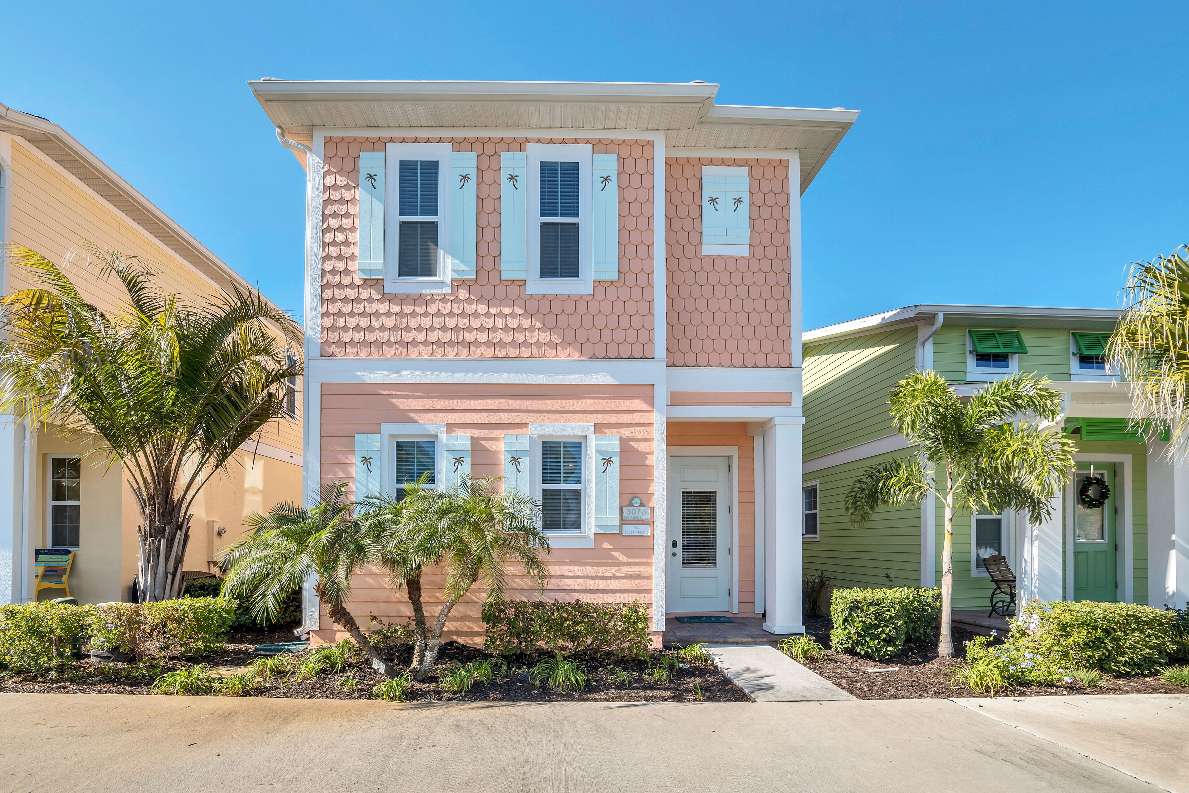 A two-story house with a pink and white exterior, a front porch, and a small garden with palm trees and shrubs.
