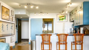 A modern kitchen with a central island, bar stools, and a view of the living area. - Summer Deal Ocean View Kailua-Kona Condo Steps to Shops and Dining (Kailua-Kona)