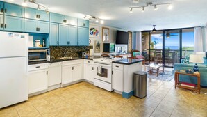 A modern kitchen with blue cabinets, white appliances, and a large island. There is a dining area with a view of the ocean. - Summer Deal Ocean View Kailua-Kona Condo Steps to Shops and Dining (Kailua-Kona)
