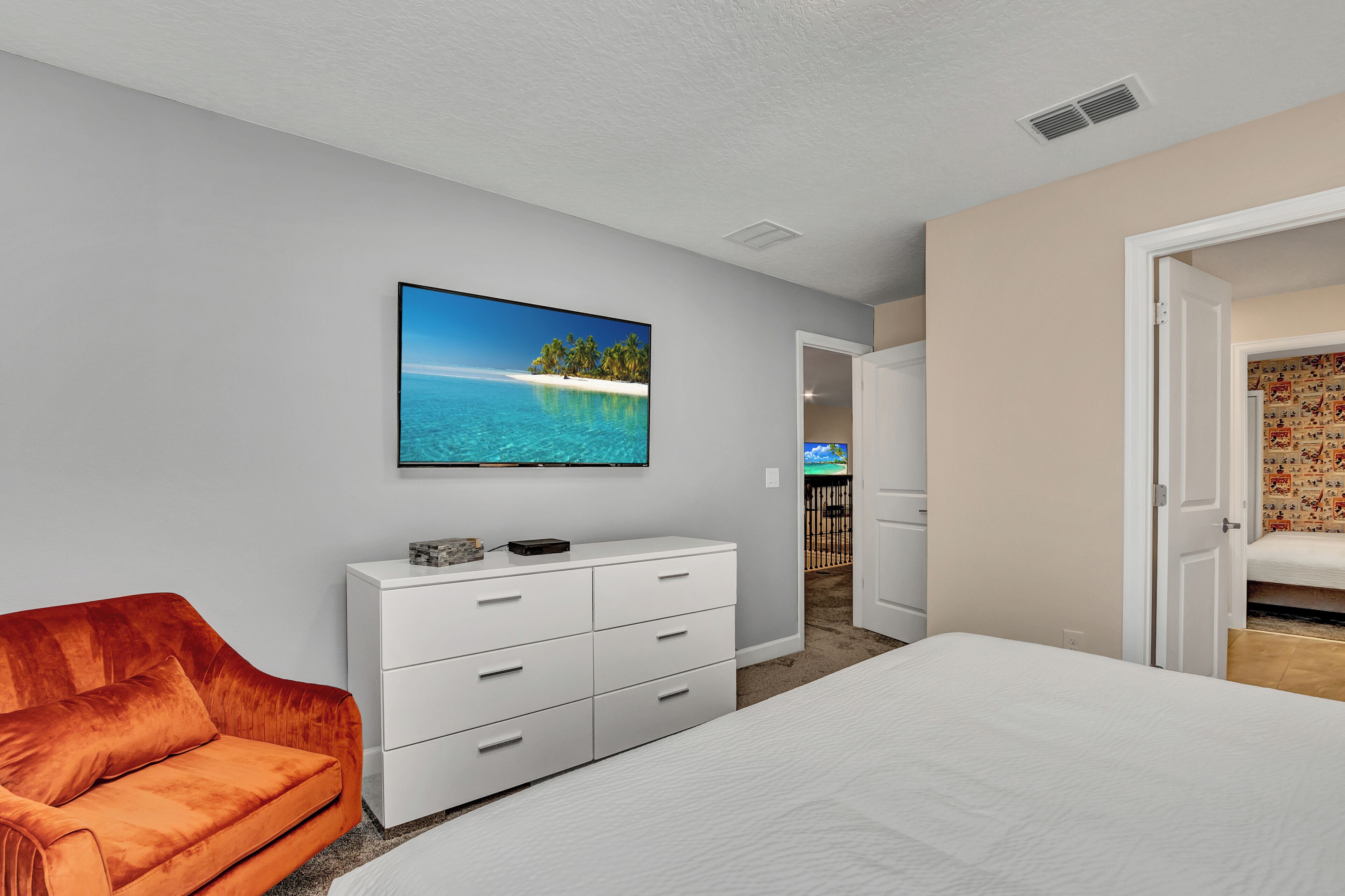 Une chambre à coucher avec un lit, une commode blanche, un canapé rouge et un téléviseur fixé au mur qui diffuse une image d’une plage tropicale.