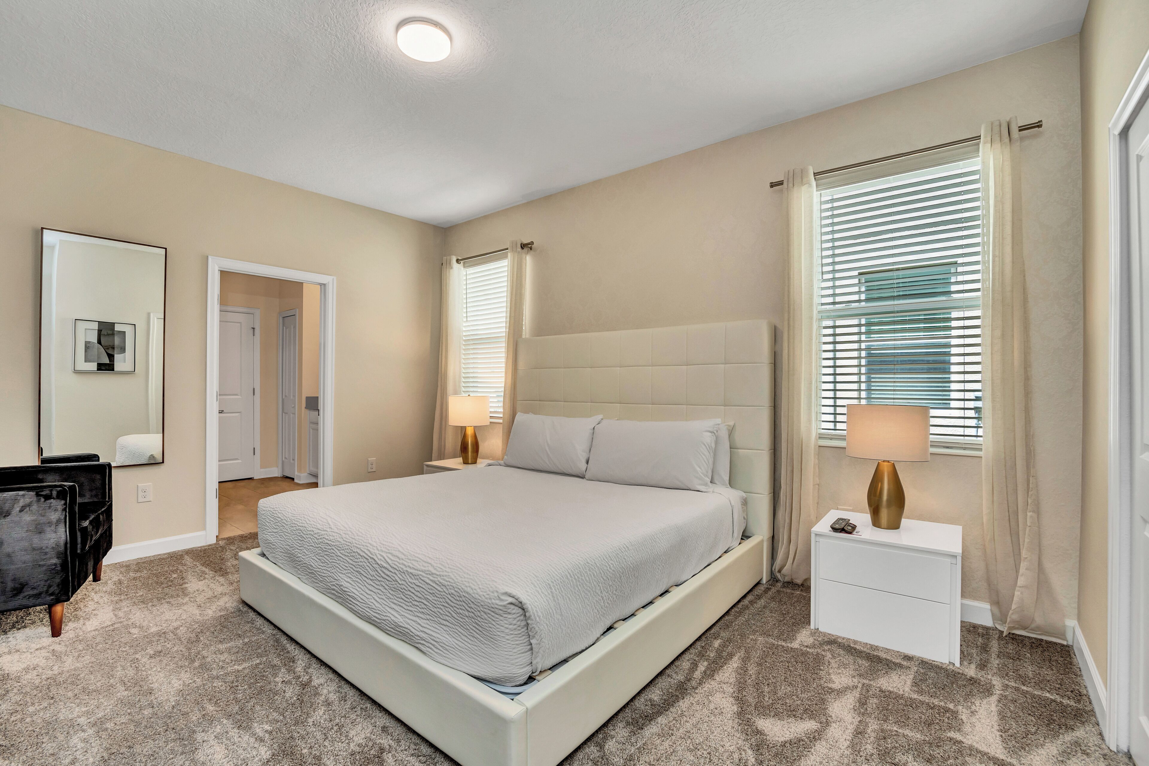 A bedroom with a large bed, a bedside table with a lamp, a dresser, a mirror, and a door leading to another room.