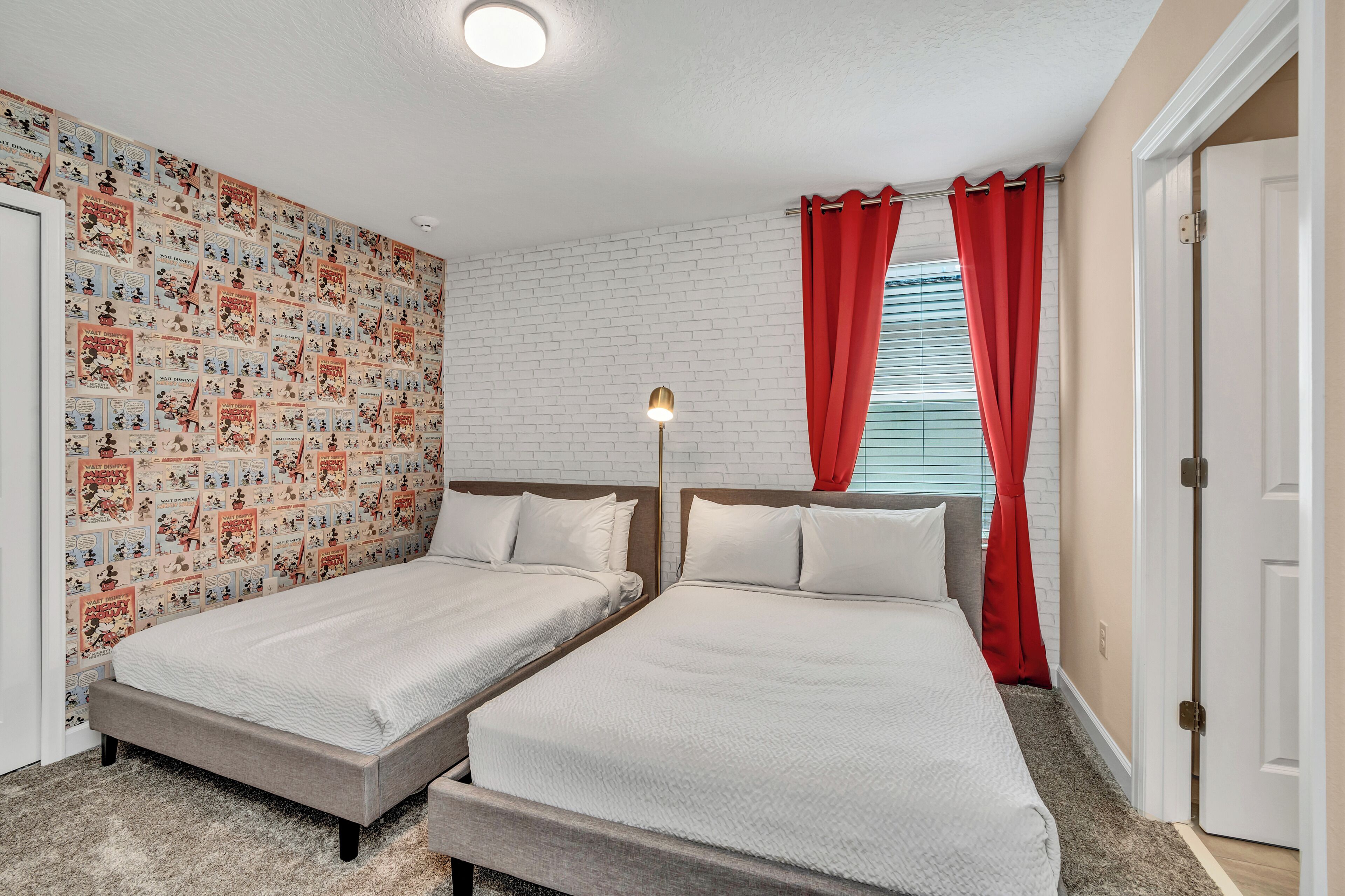 A room with two beds, a white brick wall, red curtains, and a ceiling light.