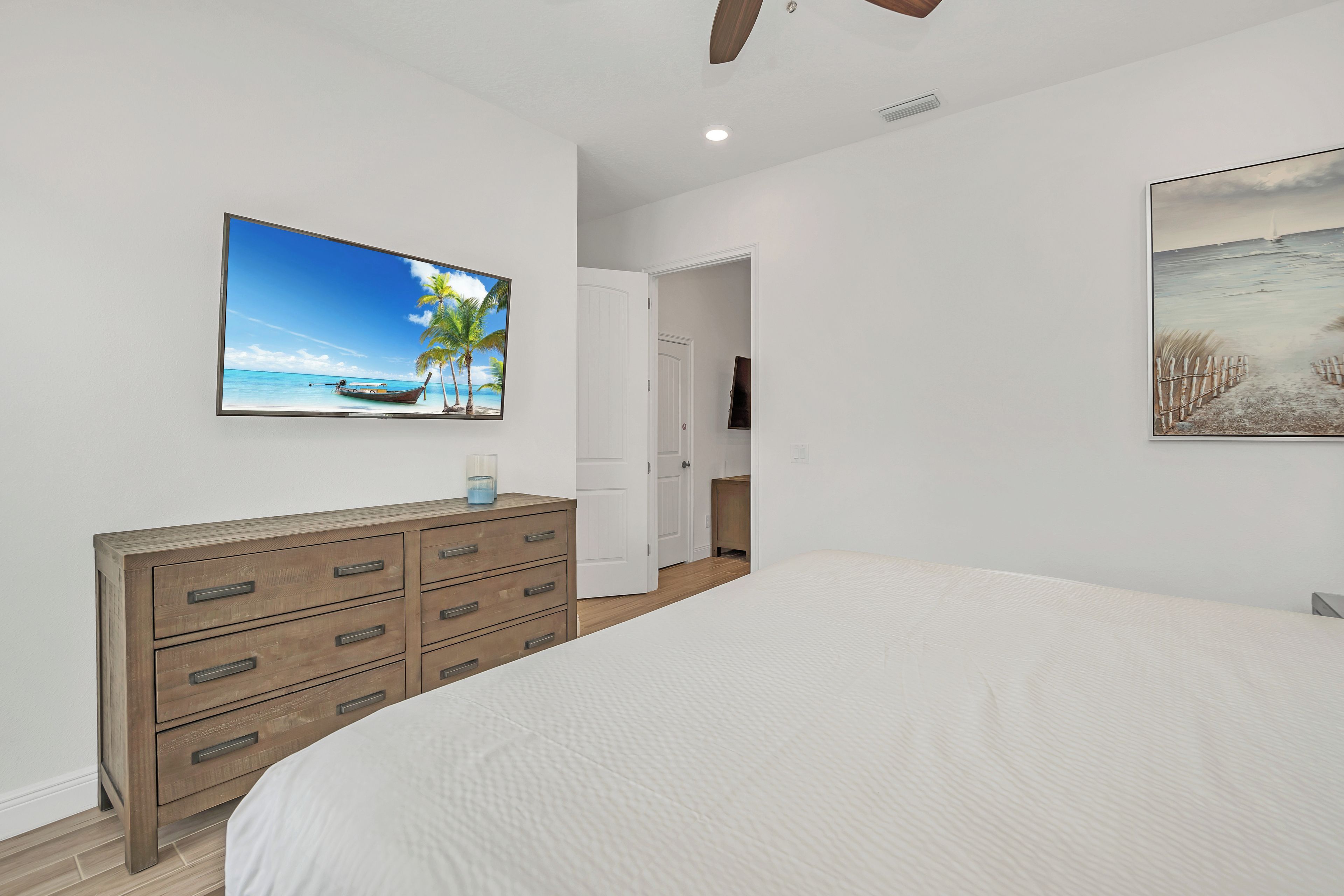 A bedroom with a large bed, a wooden dresser, a painting of a beach, and a television on the wall.
