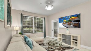 A modern living room with a beige sofa, a glass coffee table, and a painting of a tropical beach scene. - Luxurious Condo Near Disneyfree Waterpark Access (Kissimmee)