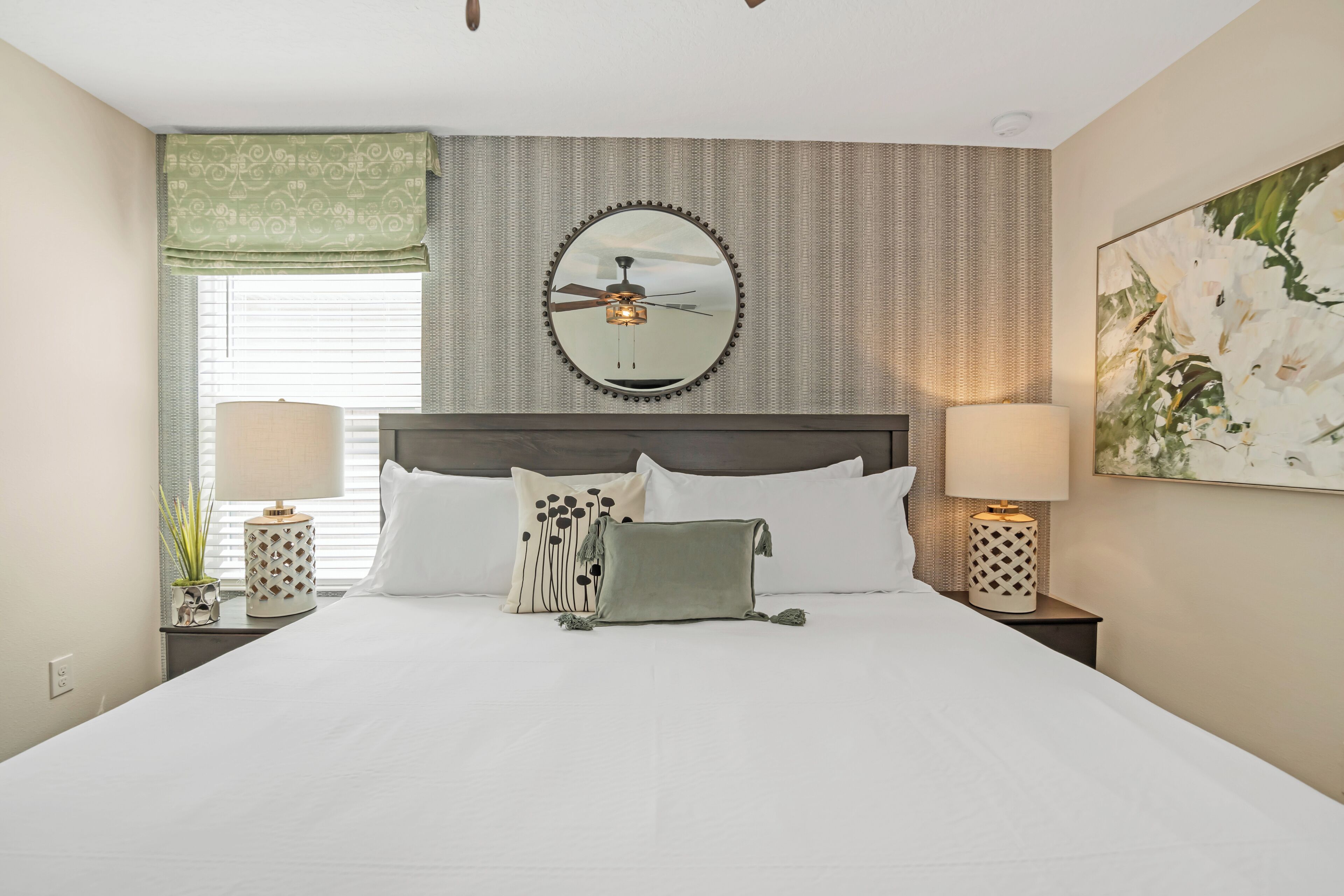 A neatly arranged hotel room with a large bed, two bedside tables with lamps, a ceiling fan, and a large floral painting on the wall.