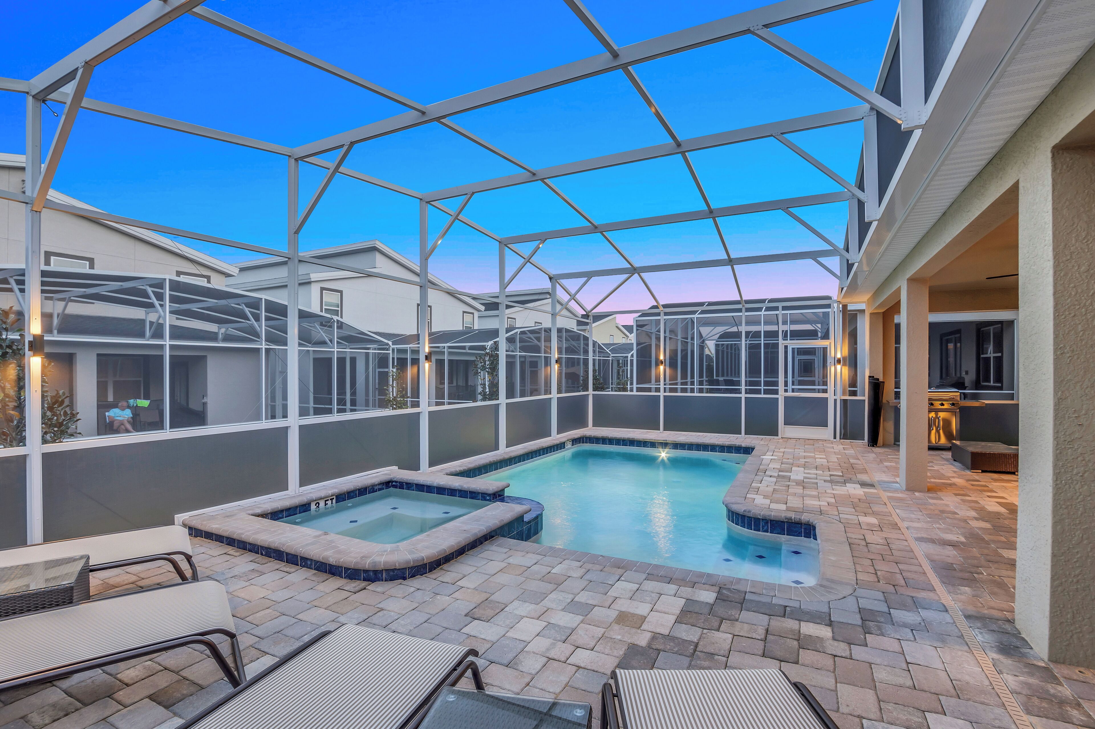 An outdoor swimming pool with a glass enclosure, surrounded by patio furniture and a brick floor.