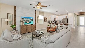 A spacious living room with a sofa, a coffee table, and a TV. There is a kitchen area with bar stools and a ceiling fan. - Large Villa, Private Pool+game Room+movie Theater! (Kissimmee)