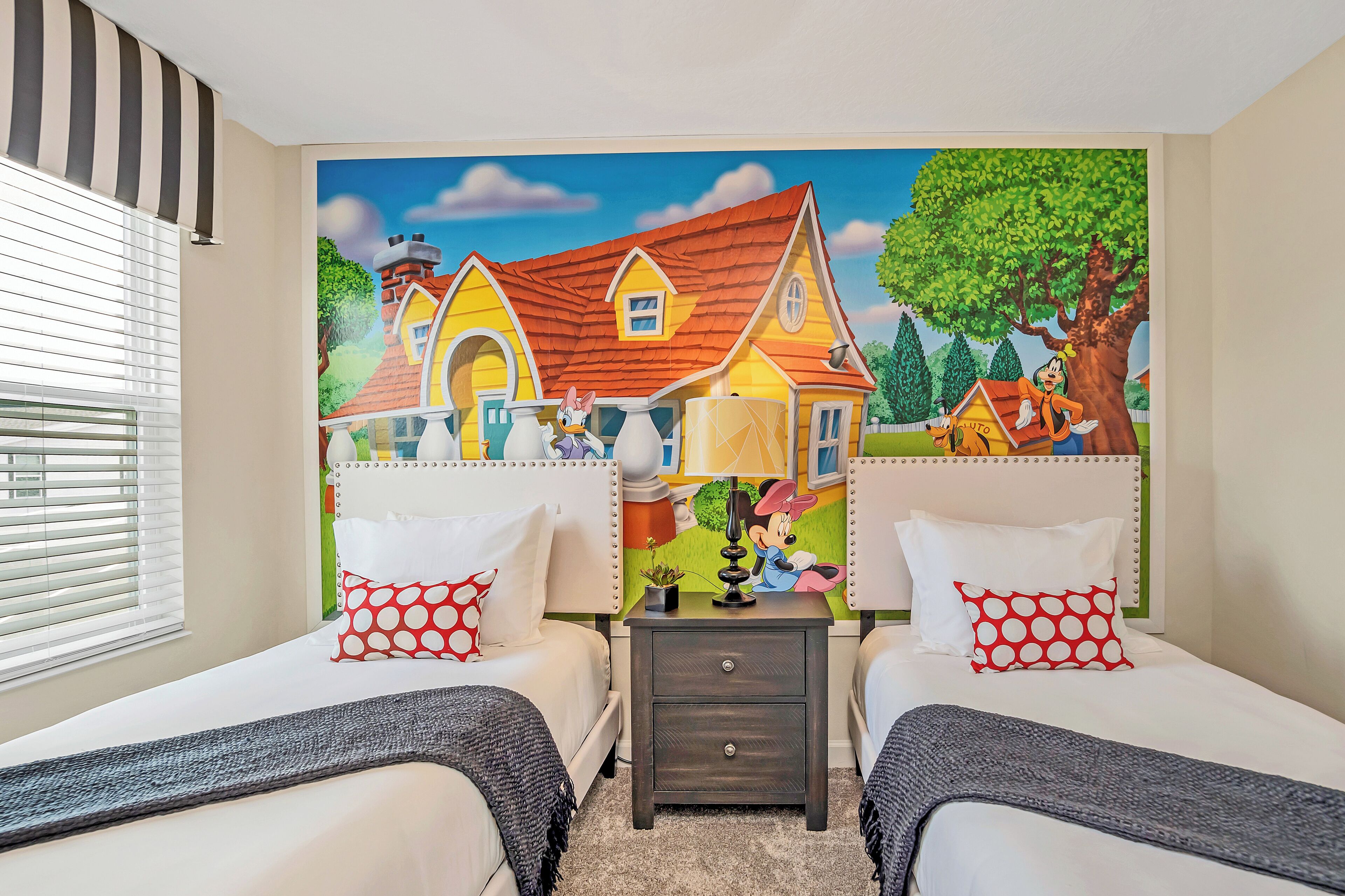 A bedroom with two beds, a nightstand, a lamp, and a mural of a house.