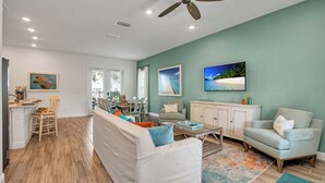 A spacious living room with a white sofa, a teal armchair, and a coffee table. There is a ceiling fan, a painting of a beach, and a television mounted on the wall. - Large Margaritaville Villa w/ Private Pool + View (Kissimmee)