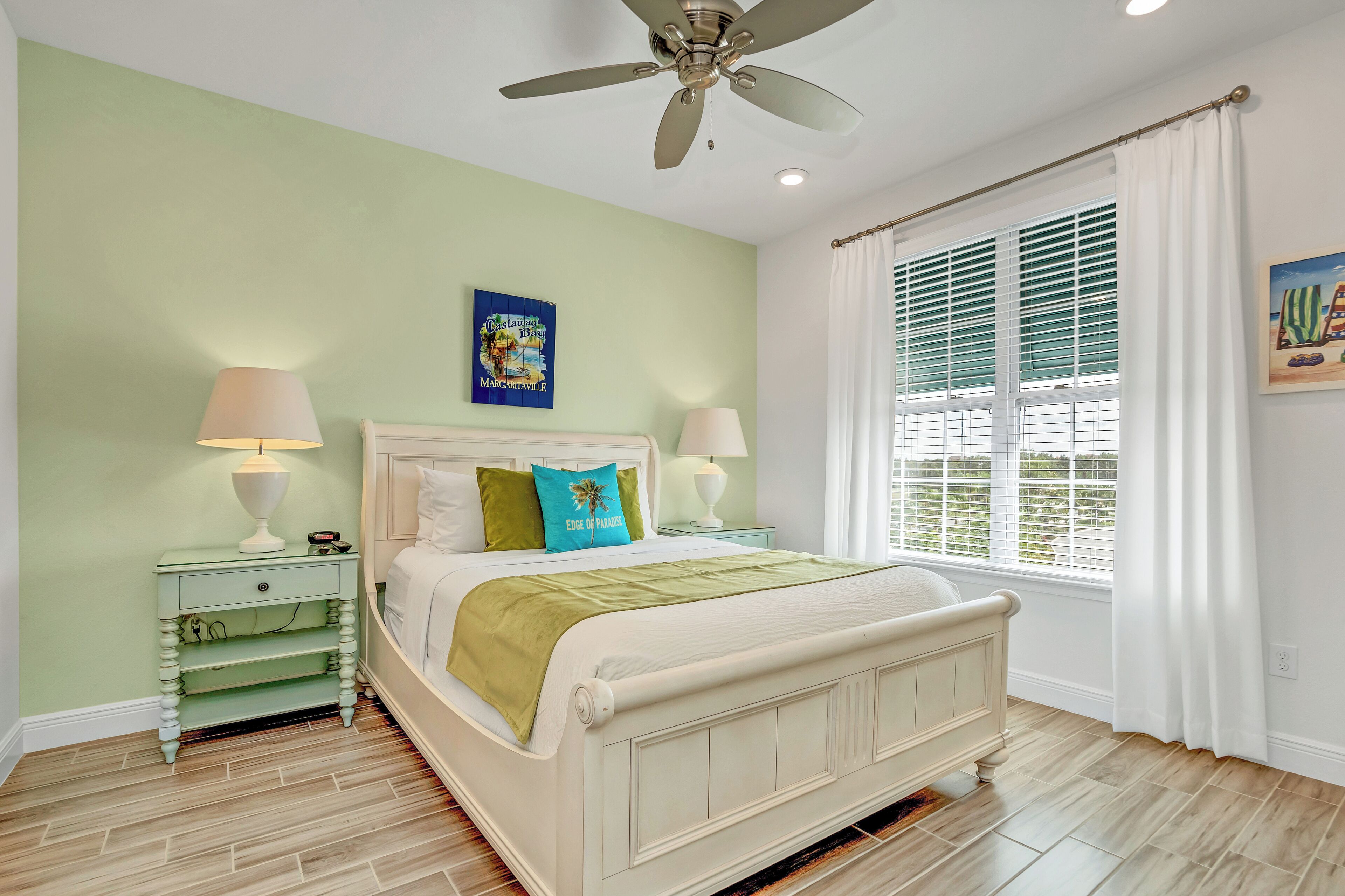 A bedroom with a bed, two bedside tables, a ceiling fan, a window with blinds, and a wall-mounted picture.