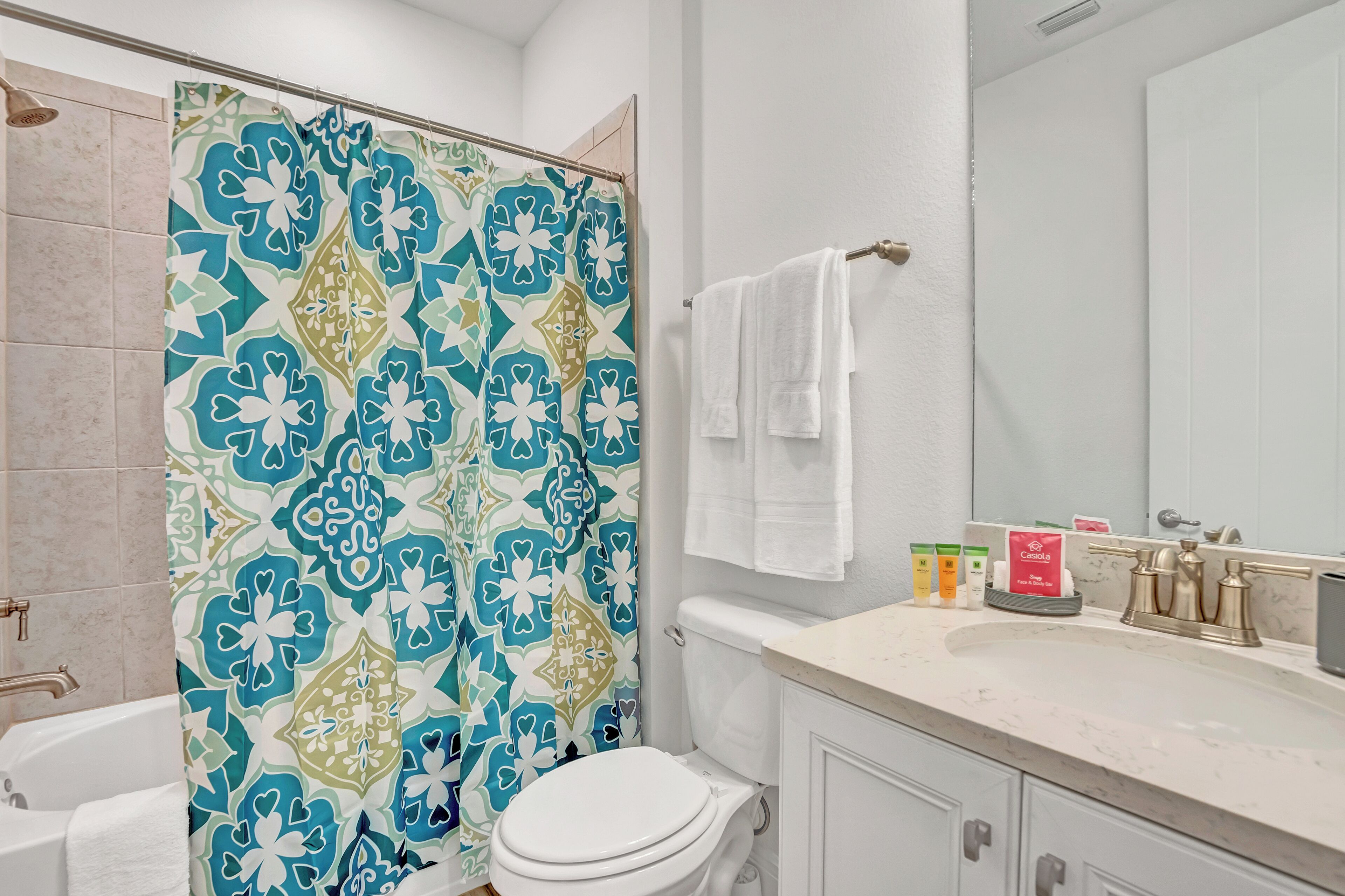 Un baño con cortina de ducha estampada, inodoro blanco, lavamanos con artículos de aseo y un espejo.