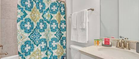 Un baño con cortina de ducha estampada, inodoro blanco, lavamanos con artículos de aseo y un espejo.