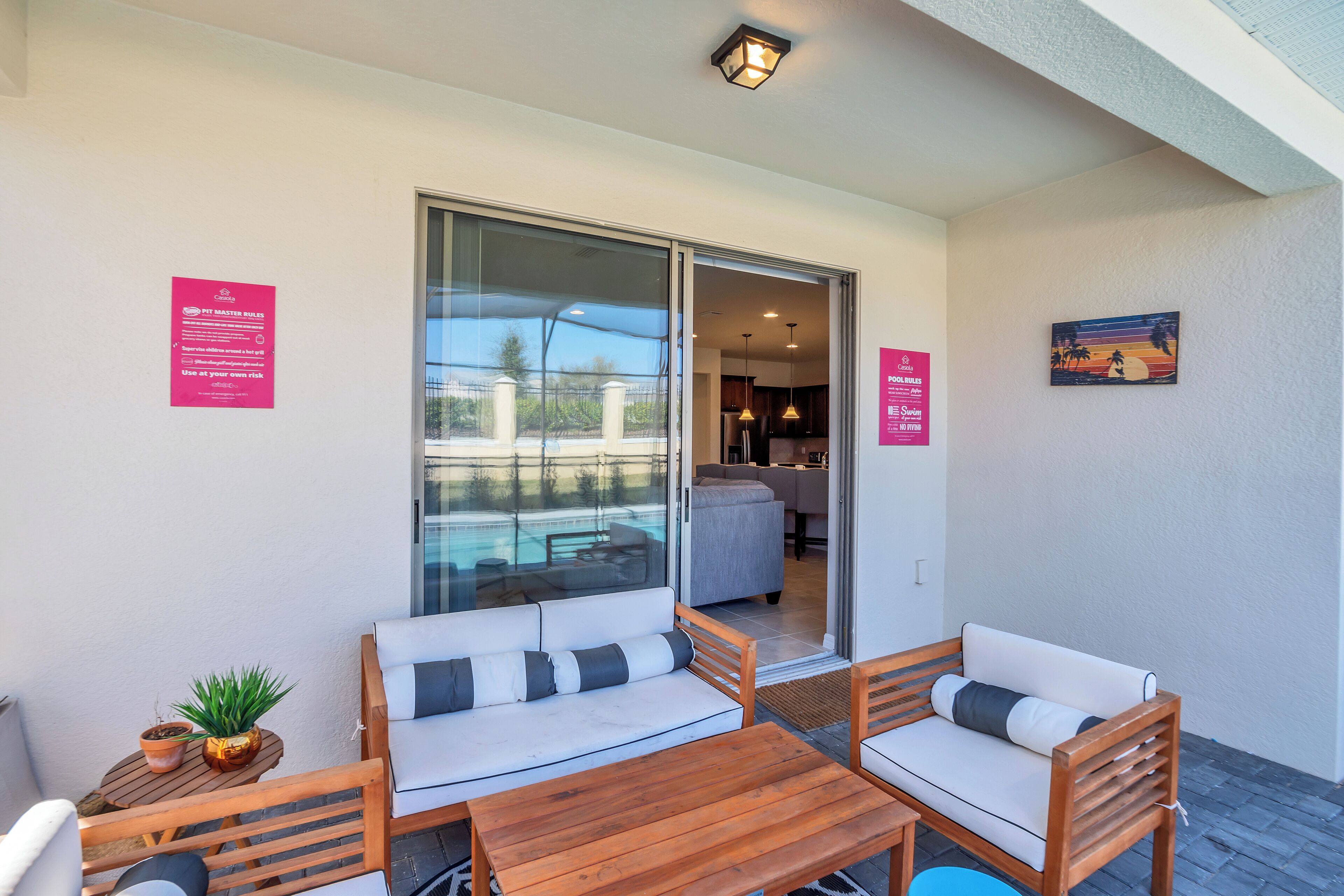 A balcony with a wooden table and two white sofas with grey cushions, a potted plant, and a glass door leading to an indoor area.