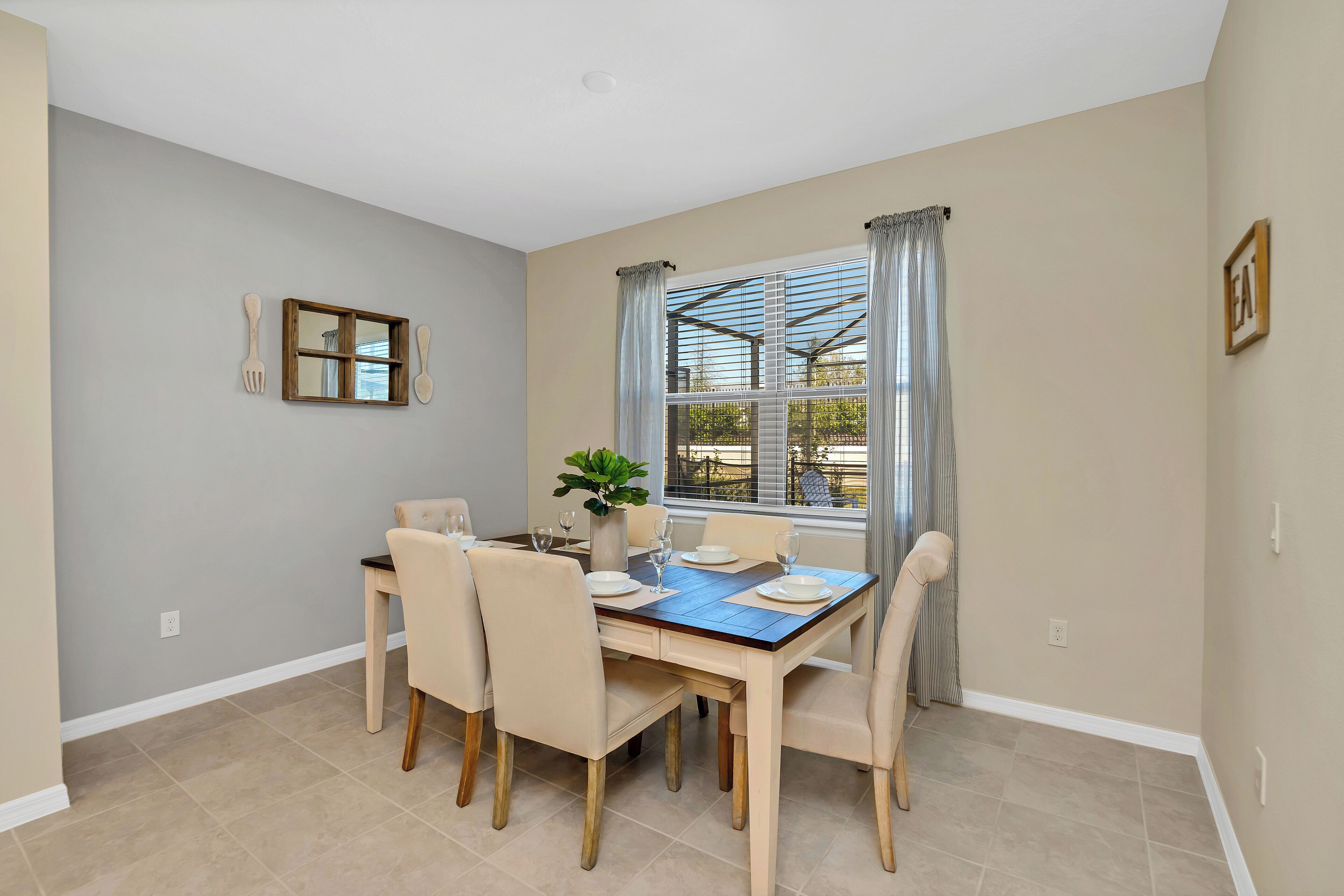 A dining area with a rectangular table set for six, beige chairs, a vase with a plant, and a window with curtains.