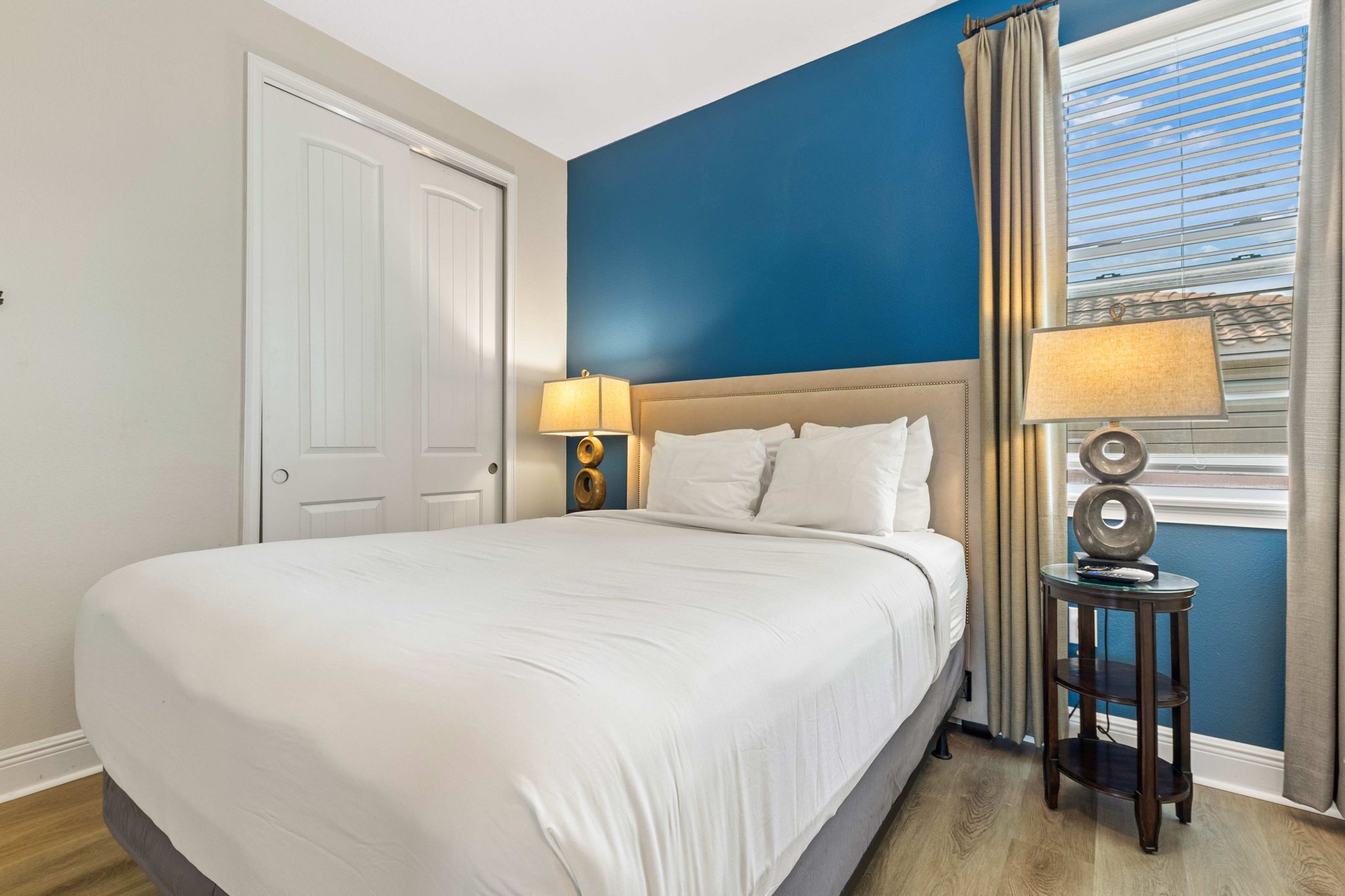 A neatly made bed with white linens in a room with blue walls and a window with blinds.