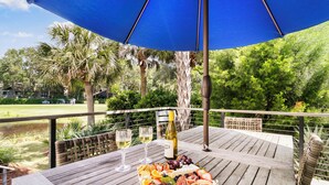 Une table en bois sur laquelle est disposée une assiette de fruits, deux verres de vin blanc et une bouteille de vin, le tout placé sous un parasol bleu sur une terrasse extérieure offrant une vue sur un terrain de golf et des palmiers.