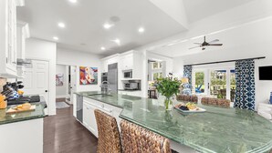 Une cuisine moderne avec des meubles blancs, un grand îlot central doté d’un plan de travail en marbre vert, des chaises en osier et un ventilateur de plafond.
