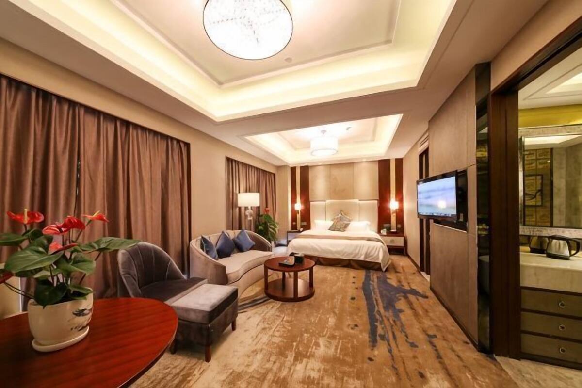 A hotel room with a bed, sofa, TV, and a dining table with a plant.