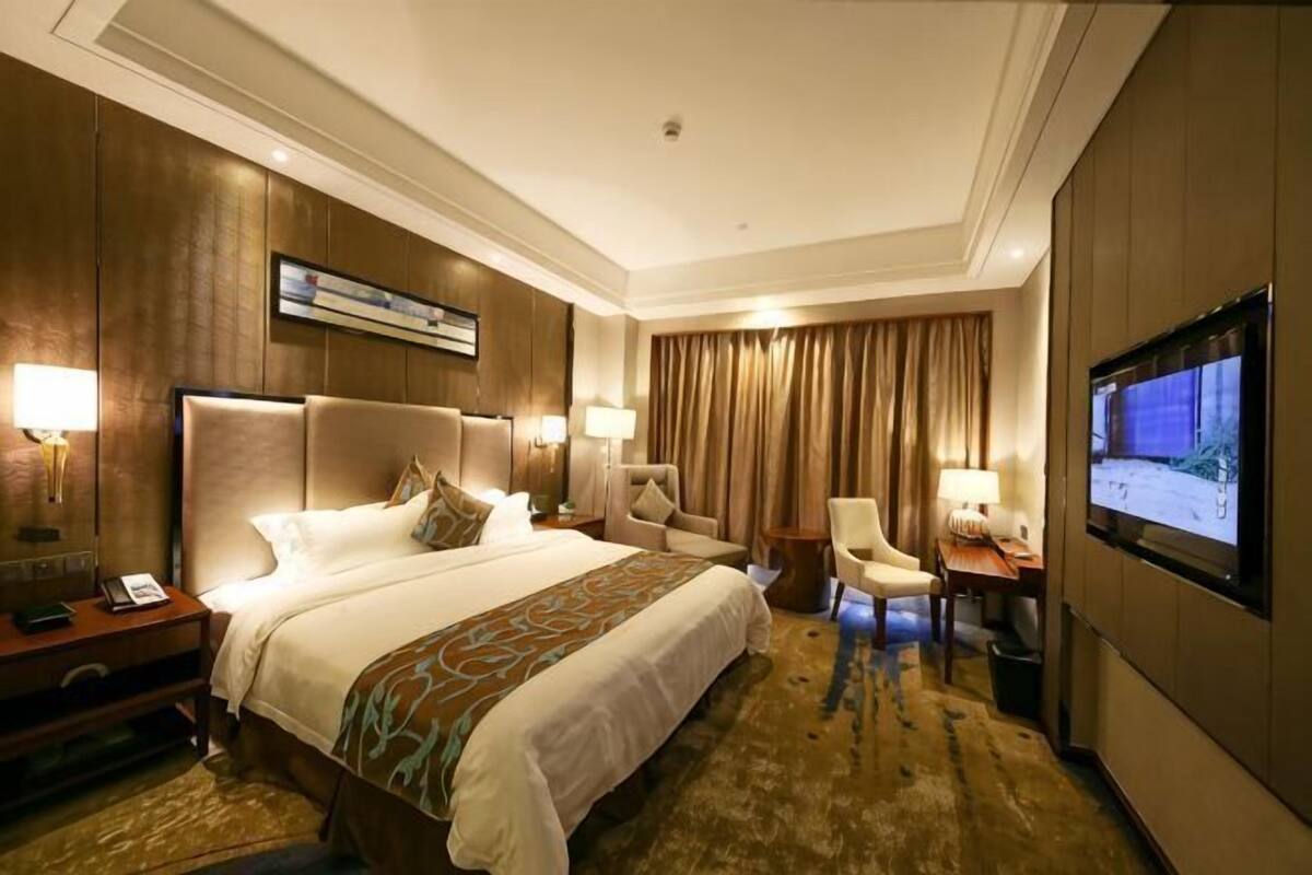 A hotel room with a large bed, two bedside tables, a desk, a chair, a TV, and a large window with curtains.