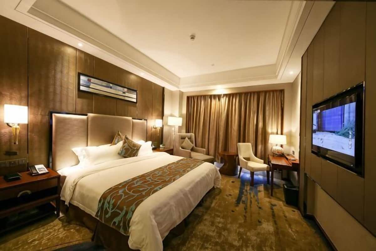 Ein Hotelzimmer mit einem großen Bett, zwei Nachttischen, einem Schreibtisch, einem Sessel, einem Fernseher und einem großen Fenster mit Vorhängen.