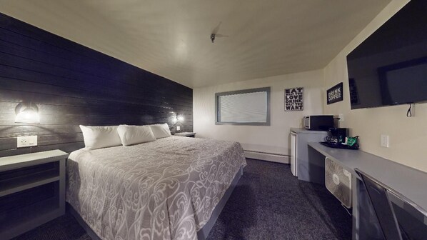 A modern hotel room with a large bed, bedside tables, a desk with a chair, a flat-screen TV, and a small kitchenette.